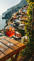 Glass of aperol spritz on a wooden table on a balcony overlooking Positano Italy on a sunny day with italian buildings in the background prompting desire to drink the glass and travel