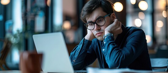 Young unhappy man office worker feeling bored at work looking at laptop with demotivated face expression while sitting at workplace in office distracted male worker feeling tired of monotonous