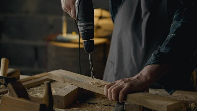 A middle-aged builder drills a hole on wood with an electric drill. In a production room. General angle. A novice technician. Works at a carpenter's table. At the workplace, hobby.