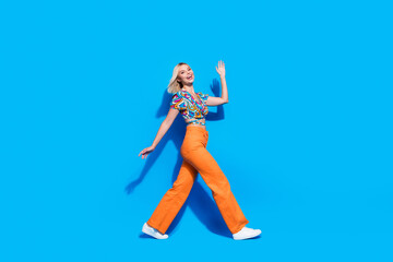 Fototapeta premium Full size profile portrait of cheerful stunning girl walking arm palm waving hi empty space isolated on blue color background