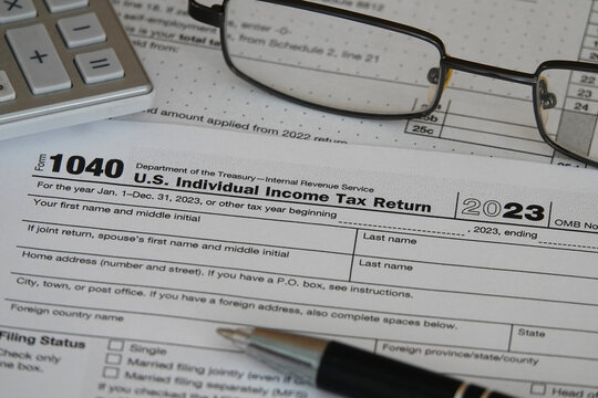 An IRS 1040 tax year 2023 form is shown in 2024, along with an ink pen, calculator, and glasses. The Internal Revenue Service tax filing deadline in 2024 is scheduled for April 15.