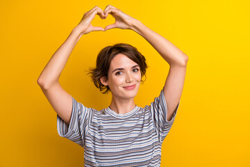 Portrait of good mood cheerful girl with short hairstyle wear grey t-shirt show heart over head isolated on yellow color background