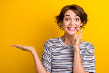 Portrait of cheerful girl with short hairstyle wear grey t-shirt palm show offer finger on lip isolated on yellow color background