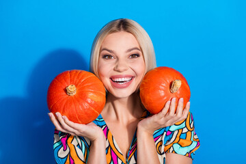 Photo of toothy beaming cute girl with bob hairdo dressed print shirt hold two squash on cheekbones isolated on blue color background