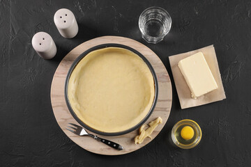 Pie tin with fresh dough and ingredients on black table, flat lay. Making quiche