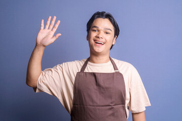 Friendly young barista wear apron waving hand saying hi or goodbye over blue background