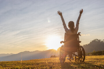 Person sitting on a wheelchair enjoying with raised hands on the mountain at the sunset time. - obrazy, fototapety, plakaty