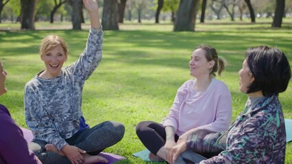 Cheerful female yoga teacher and group of mature women sitting on green grass in the park, stacking hands together and raising arms up after outdoor fitness class