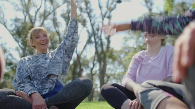 Group of jolly mature women sitting in a circle on green grass in the park, hand stacking, raising arms and happily smiling after outdoor yoga practice