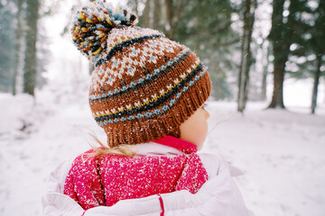 Little girl stands under snowfall in a snowy forest and looks to the side. Back view