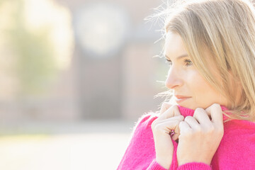 This image captures a young Caucasian woman in a vibrant pink sweater, lost in thought. The soft focus on the background bathes the scene in a warm, hazy light typical of a crisp autumn day. The woman