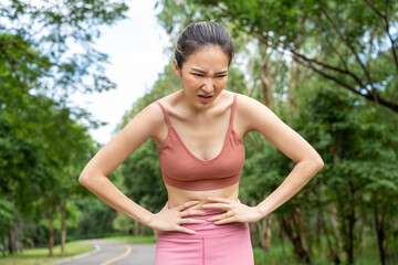 Young atractive Asian woman in fitness clothes with stomach pain putting her hands over her stomach while standing at running track of a local park