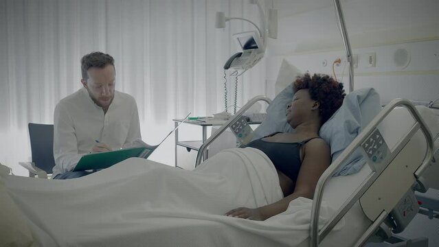Young African Woman With Curly Hair Lying in Clinic Bed Talking to Physician