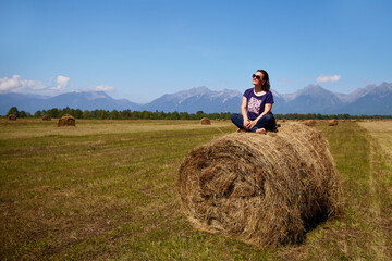 Naklejka na ściany i meble A woman is sitting on a haystack in a field with a beautiful view.