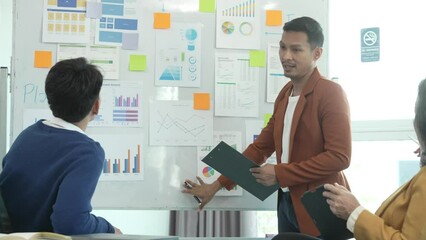 Asian business manager is leading a meeting, engaging with trainees in a bright room with data charts, indicating a monthly review or training session. - Powered by Adobe