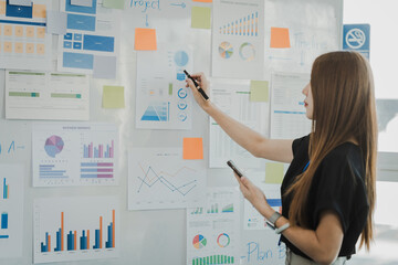 Asian female office worker presenting a business report with graphs and charts on a board, indicating a strategic or analytical discussion in a corporate environment. - obrazy, fototapety, plakaty