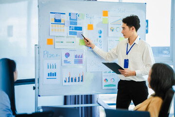 mature Asian business manager is leading a meeting, engaging with trainees in a bright room with data charts, indicating a monthly review or training session.