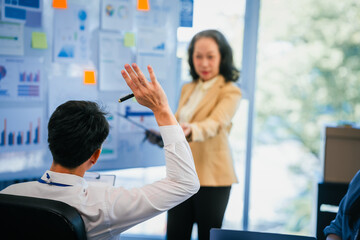 mature Asian business manager is leading a meeting, engaging with trainees in a bright room with data charts, indicating a monthly review or training session.