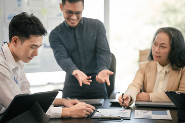 diverse group of Asian professionals, including middle-aged and mature individuals, gathered around a table in a business setting, discussing documents with focused attention.