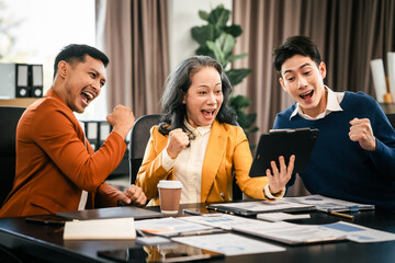 Asian colleagues are in a bright office, celebrating a moment of success. They are gathered around a tablet, with expressions of joy and achievement.