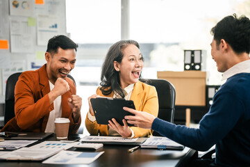 Asian colleagues are in a bright office, celebrating a moment of success. They are gathered around a tablet, with expressions of joy and achievement.
