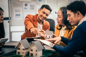 Asian real estate team engaged in a discussion, with two men and a woman focusing on a house model on a table, suggesting a planning or sales meeting. - obrazy, fototapety, plakaty