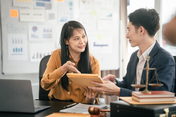 Asian man and woman collaborating professional setting, law office. reviewing documents on tablet, with laptop, contract papers, law books, and justice scale on the desk.