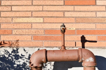 Large rusted pipe sticks out of ground against a red brick wall.