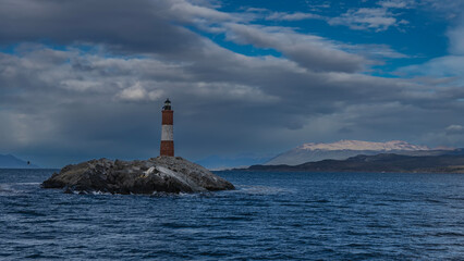 The old famous lighthouse Les Eclaireurs at the edge of the world. The red-and-white striped tower stands on a small rocky island in the Beagle Channel. Ripples on the water. Clouds in the blue sky.  