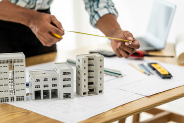 Close up Asian individual architect or engineer holding building scale model of multi-story building. design or review of architectural plans laid out on work surface in front of them.