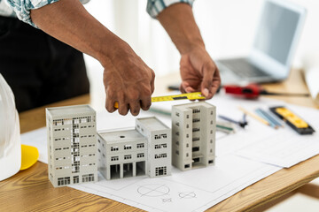 Close up Asian individual architect or engineer holding building scale model of multi-story building. design or review of architectural plans laid out on work surface in front of them.