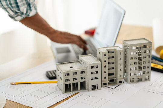 Close up Asian individual architect or engineer holding building scale model of multi-story building. design or review of architectural plans laid out on work surface in front of them.