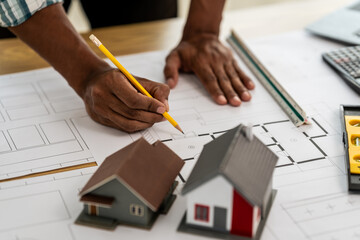 Asian person hands working on architectural blueprints with a pencil, alongside scale models of...