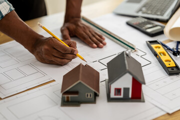 Asian person hands working on architectural blueprints with a pencil, alongside scale models of houses, a laptop, and construction tools on the desk.