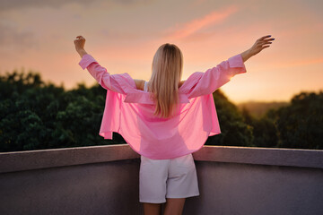 Back view of a woman standing with arms outstretched, embracing a colorful sunset