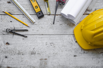 Top view construction tools such as a yellow hard hat, spirit level, measuring tape, folding ruler arrayed against a wooden plank background.