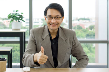 A man wearing a formal suit smiles and shows a thumbs up sign with his hand to express approval.