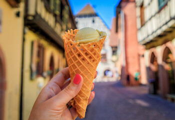 Woman s hand holding artisanal pistachio gelato in a cone, with half timbered in Kaysersberg, a popular village on the Alsatian Wine Route in France