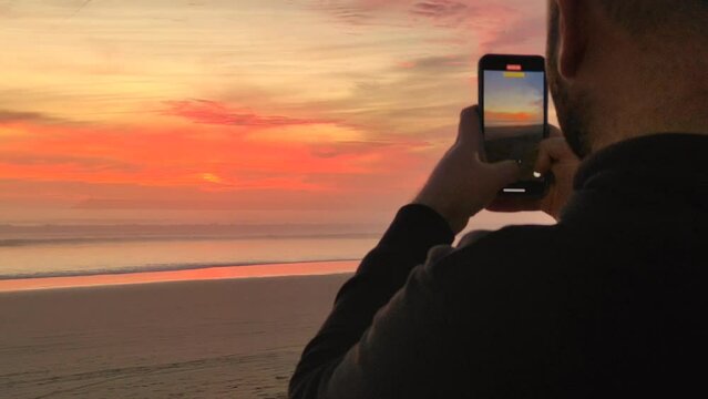 Young man male tourist takes pictures, shoots video of sunset on beach on modern smartphone. Beautiful bright sunset on the sea, ocean. boy enjoys nature, dreams, relaxes. Hands with phone