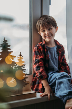 Smiling boy sitting on window sill near Christmas decoration at home