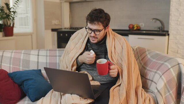 Male with eyeglasses talks on laptop, facing flu at home. Wrapped in a duvet, affected by the cold