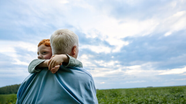 Grandfather Carrying Grandson In Field Under Cloudy Sky At Sunset