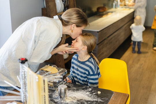 Mother kissing daughter on forehead near pasta maker at dining table