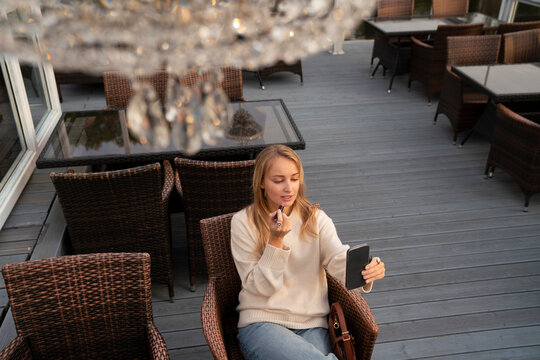 Young woman looking at smart phone and applying lipstick in cafe