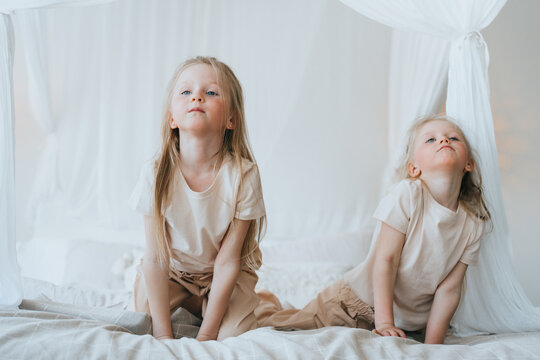 Two adorable sisters playing and making faces on a white bed, with sheer curtains creating a whimsical, dreamy bedroom atmosphere - Powered by Adobe