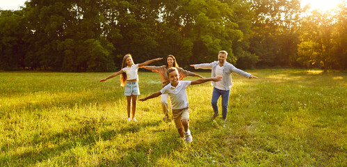 Naklejka na ściany i meble Happy family running and 'playing plane having fun in summer park enjoying sunny day in nature. Mother, father and their children boy and girl walking on green grass outdoors at sunset together.