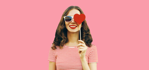 Portrait of beautiful young woman with red heart shaped lollipop wearing sunglasses on pink studio...