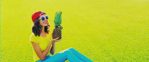 Summer portrait of happy smiling young woman with pineapple and sunglasses sitting on the grass in summer park