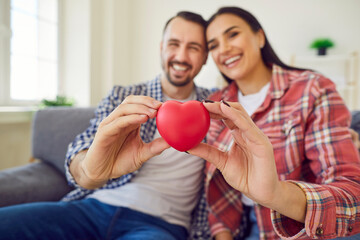 Happy couple holding a red heart in their hands. Cheerful, smiling man and woman sitting on the couch and holding a symbolic heart. Close up of a red plastic heart. Love, affection, health concept - Powered by Adobe