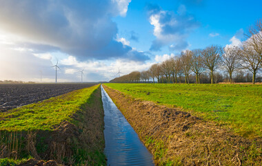 Wind turbines on the horizon of an agricultural field along a ditch in bright sunlight in winter,...